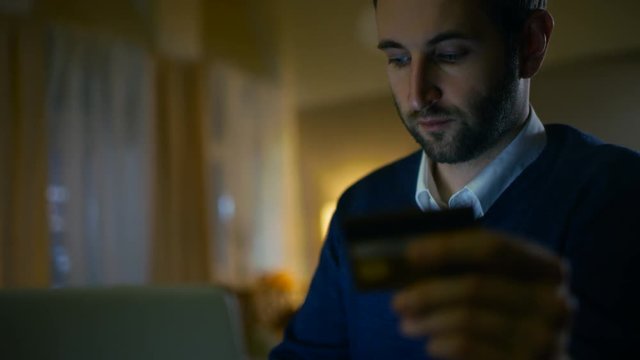 Close-up of a Man Sitting at His Desk with a Laptop Holding Credit Card and Making a Purchase from the Internet. His Apartment Has Skyscrapers View. Shot on RED EPIC-W 8K Helium Cinema Camera.
