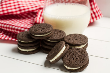 Vaso de Leche con galletas de chocolate rellenas sobre una bandeja blanca y un paño a cuadros de...