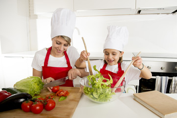 mother and little daughter cooking together with hat apron preparing salad at home kitchen