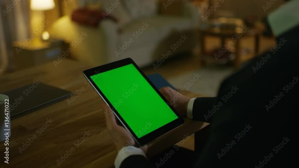 Poster at home man sits at his desk and holds tablet computer with green screen on it. his apartment is don
