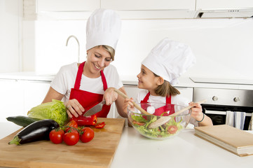 mother and little daughter cooking together with hat apron preparing salad at home kitchen