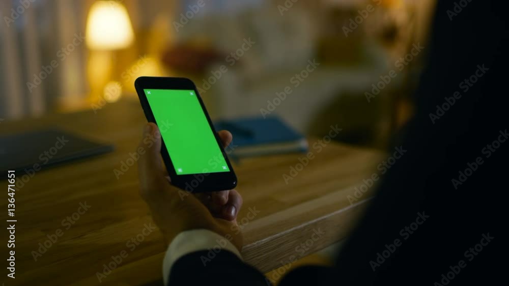Poster at home man sits at his desk and uses smartphone with green screen on it. he drinks from a cup. his 