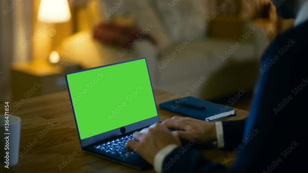 Poster at home man sits at his desk and types on a laptop with green screen on it. his apartment is done in
