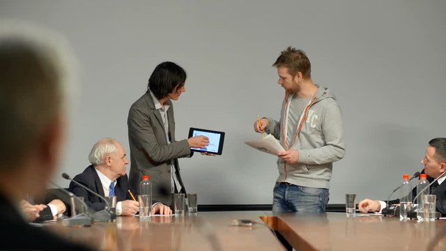 Two young male collegues and friends from new IT startup stay opposite group of investors. Students make presentation of unique internet service application to get further investments from Board of