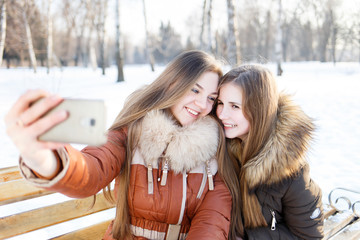 Two smiling girls make selfie on bench in winter park