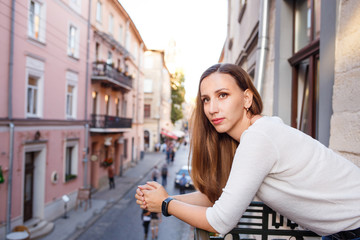 Fototapeta na wymiar Young beautiful woman standing on the balcony and looking out on street of european city