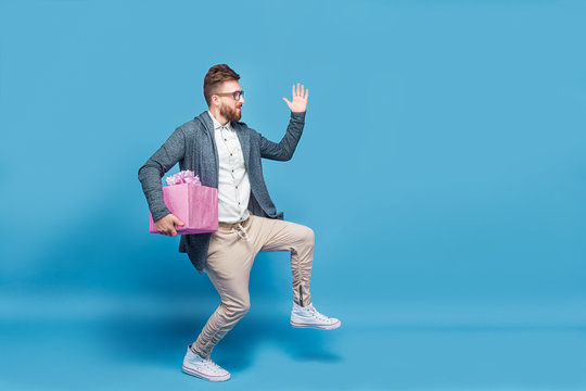 Young Man In Casual Clothing Creeping Secretly With Pink Gift Box In Hand On Blue Studio Background.