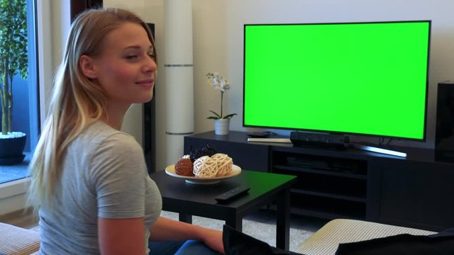 A young, beautiful woman sits on a couch in a living room and watches a TV with a green screen, then turns to the camera and nods with a smile