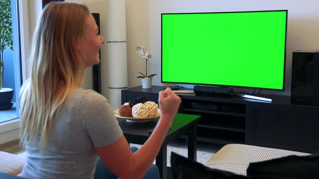 A blonde woman sits on a couch in a living room, watches a TV with a green screen and celebrates