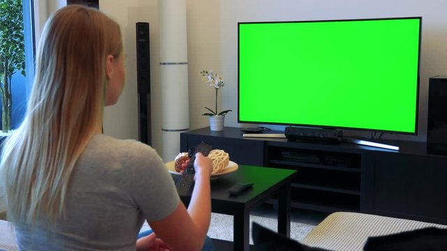 A blonde woman sits on a couch in a living room, watches a TV with a green screen and uses a TV controller