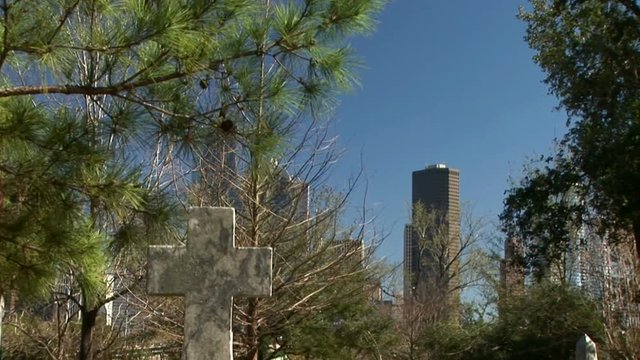 Camera pans down from large office buildings in the background to a cross shaped headstone.