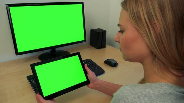 A young woman sits at a desk and looks in turns at a computer screen and a tablet (both green screen)