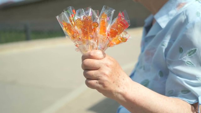 Senior adult woman selling homemade sugar lollipops in individual packaging in summer street while passers going by sidewalk