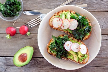 Avocado toast with kale and radish on whole grain bread, overhead scene on a rustic wood background