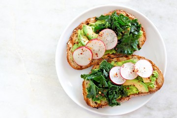 Plate of avocado toast with kale and radish on whole grain bread, top view on a marble background