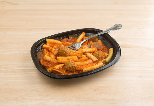 Rigatoni pasta with meatballs and sausage TV dinner with a fork in the food atop a wood table. 