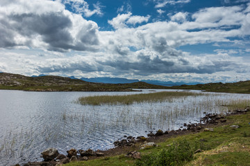 Fototapeta premium Assynt Peninsula, Scotland - June 7, 2012: Loch Sgeireach is a lake along B869. Here under blue sky with large white clouds. Reet in water, some grass upfront. Horizon of mountains.