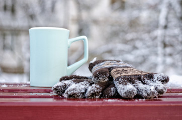 Blue a mug and women's knitted gloves on a bench in the winter, the trees in the background. Close-up