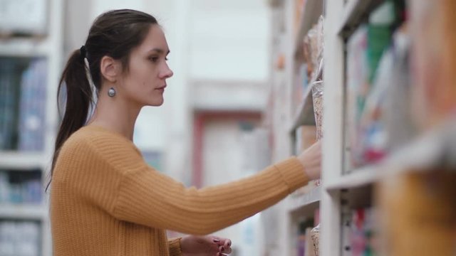 Young beautiful woman standing in front of shelves with groats, holding one and try choose something in supermarket. 4K