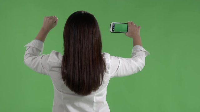 Rear view of woman with raised fist taking pictures or recording video with her camera phone against a green screen