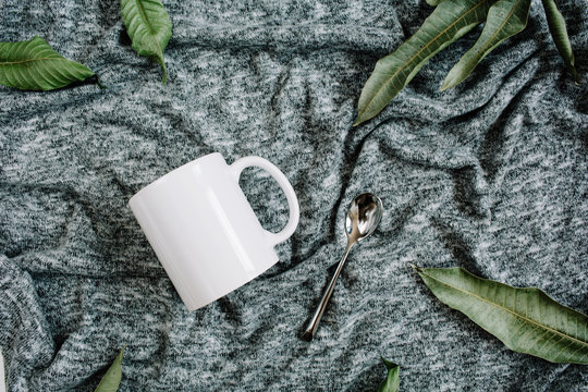 Blank template of white mug, spoon and green leaves on grey textile background. Flat lay, top view.