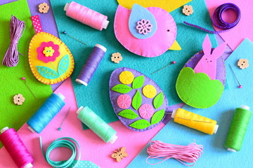 Fototapeta na wymiar Felt Easter eggs with flowers and bunny, a felt bird. Easter ornaments set, colored thread spools, felt sheets, pins, ribbons, wooden buttons on a table. Colorful Easter background. Top view. Closeup