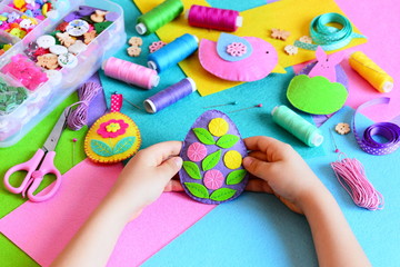 Child made a felt Easter egg ornament. Small child holds a felt Easter egg ornament in his hands. Easter crafts, craft tools and materials on a table. Festive spring crafts concept