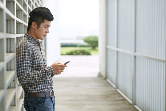 Profile View Of Young Asian Man In Jeans And Checked Shirt Standing Outdoors And Playing Games On His Smartphone