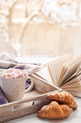Coffee cup, open book, marshmallow, croissants and plaid on a window sill in winter. Concept warm and cozy home decor. Winter breakfast. Vintage style