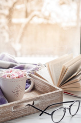 Coffee cup, open book, glasses, marshmallow and plaid on a window sill in winter. Concept warm and...