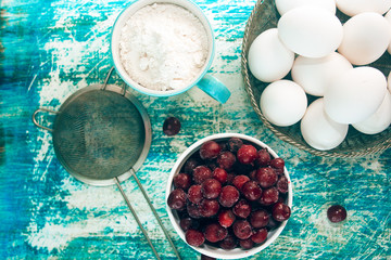 set of cooking cherry pie: a Cup of flour, a sieve, eggs, and frozen cherries in a white bowl on old rustic blue table a top view