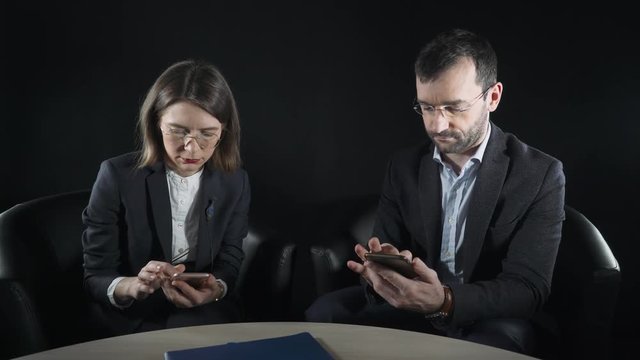 Business people sitting at the table, look in the phone and talk.  Business couple. 4k footage. Close-up. Black background.