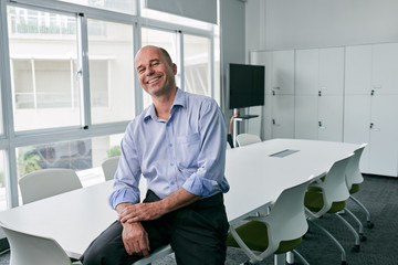 Bald middle-aged businessman sitting on office table corner and looking at camera with wide smile in meeting room