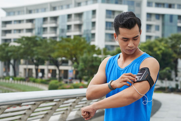 Waist-up portrait of sweaty young athlete listening to music in headphones and choosing necessary tunes for training on his smartphone