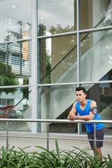 Confident young sportsman with smartphone and fitness tracker looking away seriously while standing against skyscraper window