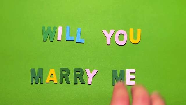 Wood characters of different colors arranged in a word will you marry me on a green background with hand slide

