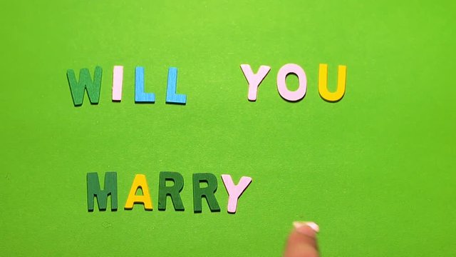 Wood characters of different colors arranged in a word will you marry me on a green background with hand slide
