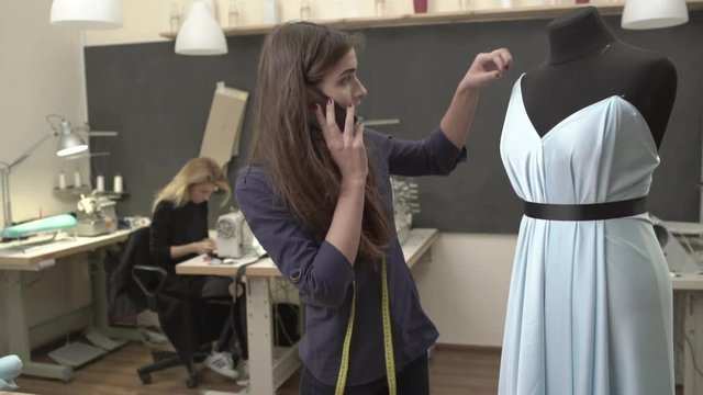 Busy Caucasian beautiful female in dark blue shirt talking on mobile phone in sewing classroom standing near mannequin with azure dress while blonde woman working at table with machine in slowmotion