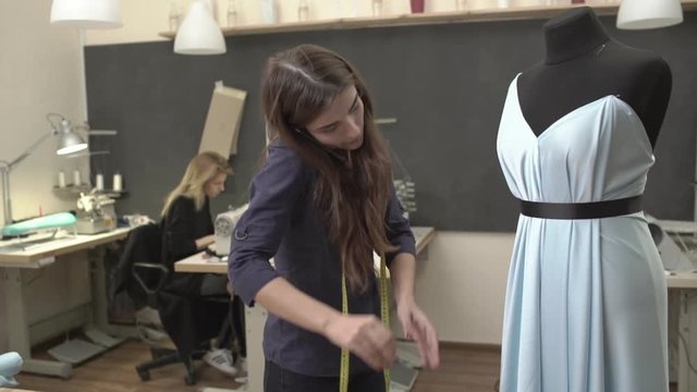 Attractive Caucasian female with long brown hair and dark blue shirt standing near mannequin with azure dress pinning it while blonde woman working at table with machine in slowmotion