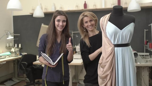 Blonde Caucasian female working on mannequin with azure dress and peach-colored cloak with woman in dark blue shirt with long brown hair standing near smiling showing ok in slowmotion