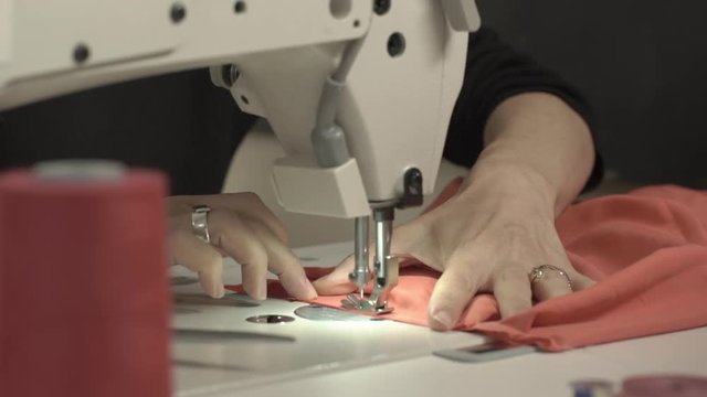 Close up footage of sewing machine stitching piece of peach-colored fabric while woman's hands holding it on table in slowmotion