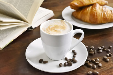 Breakfast scene with fresh croissants, coffee cup, coffee beans and open book. Relaxation morning scene with hot coffee.