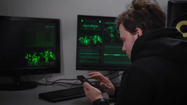 Adult male Hacker, with stubble on his face, wearing a black sports jacket sits in a secret bunker and makes cyber attacks. On your smartphone, he said the data