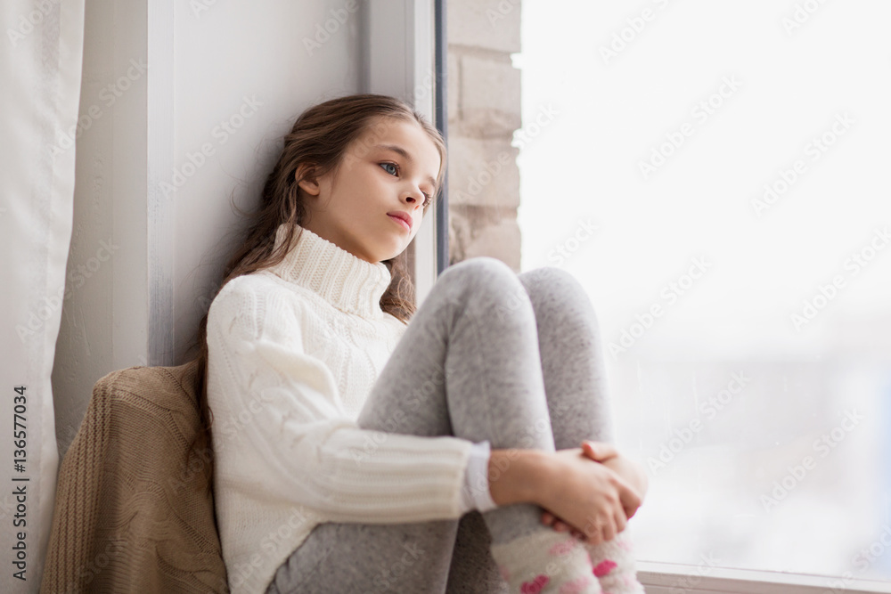 Wall mural sad girl sitting on sill at home window in winter