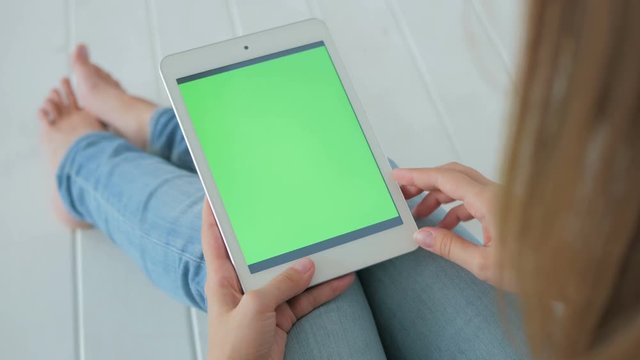 Woman sitting on the white wooden floor and using vertical tablet computer with green screen. Close up shot of woman's hands with pad. Technology and internet concept