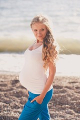 Smiling pregnant woman 20-24 year old walking at beach wearing casual white top and blue sporty pants outdoors. Looking away. Motherhood. 20s.