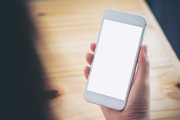 Mockup image of hand holding white mobile phone with blank white screen on vintage wood table in cafe