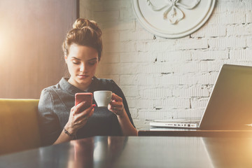 Young woman sitting in cafe at table, drinking coffee and using smartphone. On table is laptop. On...