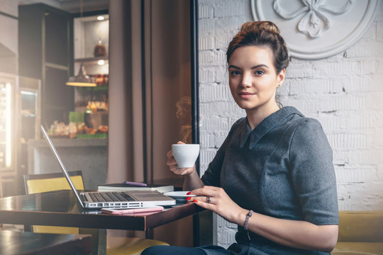 Young businesswoman sitting at table in cafe, drinking coffee and looking at camera.On table is laptop, mobile phone and notebook. Girl waiting for friends, blogging.Freelancer working outside office.