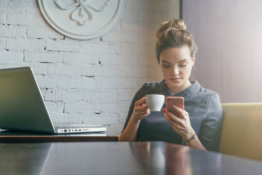 Young businesswoman sitting in cafe at table, drinking coffee and using smartphone. On table is laptop. On background white brick wall. Student learning online. Girl shopping online, checking email.